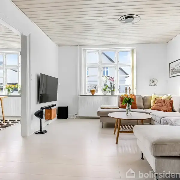 Stue med lys sofa og sofabord, dekoreret med blomster i vase. Fladskærms-TV på væggen. Store vinduer giver sollys. Spisestue i baggrunden med træmøbel og mønstret tæppe.