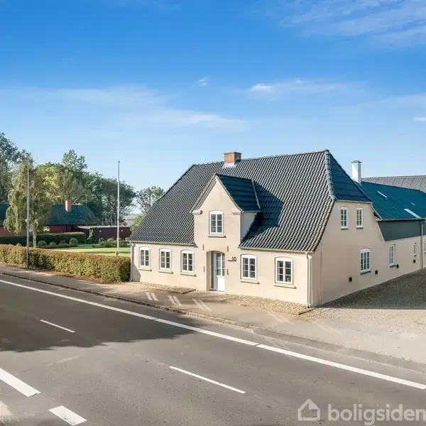 En lys beige husbygning med sort tag står stille ved en landevej. Omkringliggende er der grønne træer og en klar blå himmel.