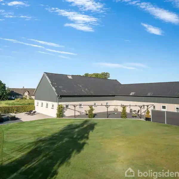 En stor, lys bygning med sort tag står på en velplejet plæne. Træer i baggrunden og en klar blå himmel skaber en fredelig landlig atmosfære.