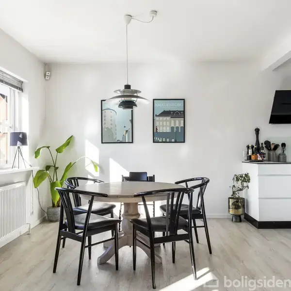 Et spisebord omgivet af sorte stole står i et lyst køkken med hvide skabe og en moderne sort emhætte. En plante og kunstværker pryder den solbelyste væg.