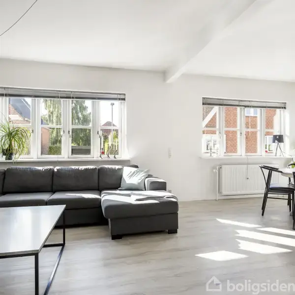 En grå hjørnesofa med puder står op ad væggen under to store vinduer. Ved siden af er et spisebord med stole. Rummet er lyst og rummeligt.