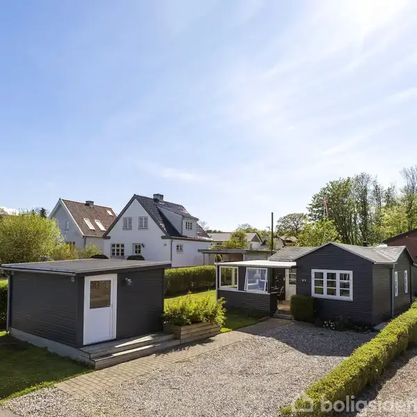 En række små huse står stille i en solbeskinnet boligkvarter. Häkkede haver omgiver husene, og en klar blå himmel strækker sig over den grønne, skovrige baggrund.