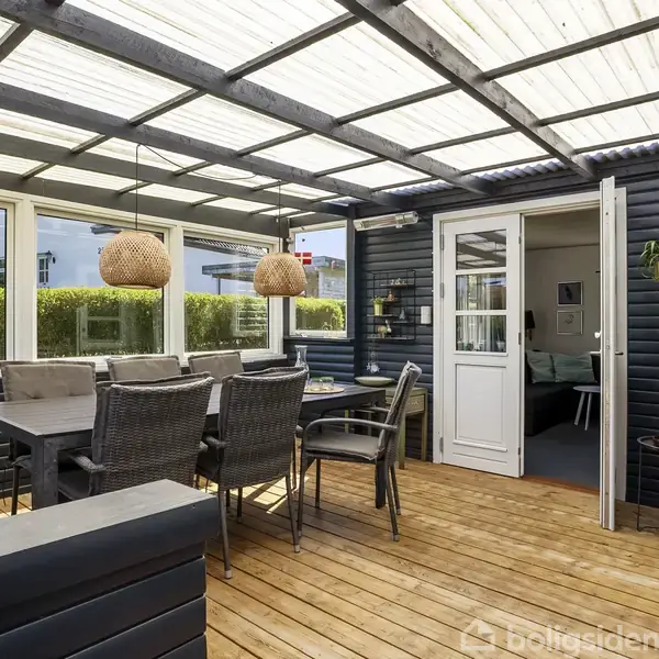 Overdækket terrasse med trægulv; spisebord med seks stole. To hængelamper over bordet. Mørk træbeklædning på vægge. Dør fører ind til huset. Grønt hegn udenfor.
