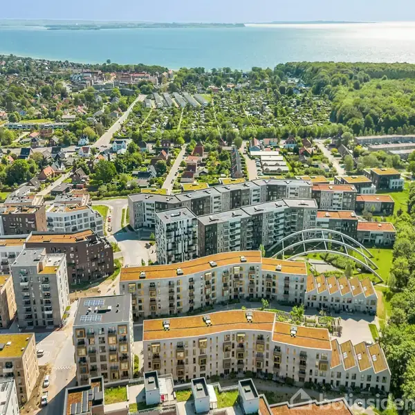 Et boligområde med moderne lejlighedskomplekser og grønne tage strækker sig gennem billedet. I baggrunden ses bylandskab og skov, som strækker sig mod kysten med klart blåt hav.