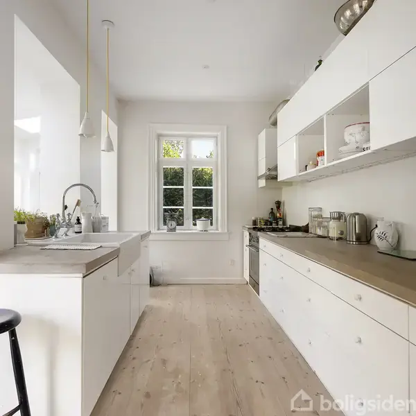 Et lyst, moderne køkken med hvid indretning og trægulv. Langs den ene væg står et bord med indbygget ovn og mikroovn. Et stort vindue giver naturligt lys. En sort skammel står ved vasken.