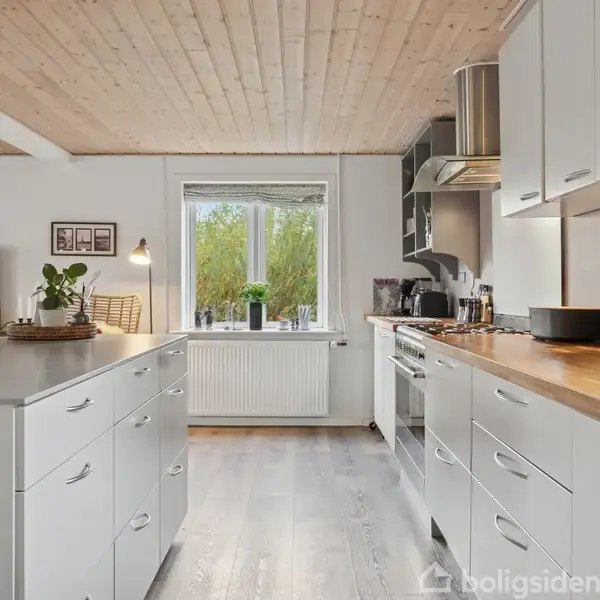 Køkken med hvide skabe og træbordplader dominerer rummet. Køleskabet og ovnen er indbygget. Et vindue åbner mod grønt udenfor. En køkkenø står centralt med planter og lamper.