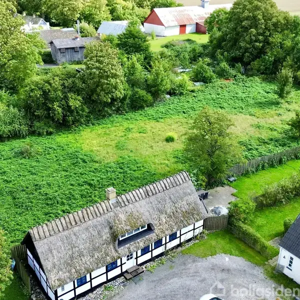En bindingsværksejendom med stråtag står stille i et frodigt, grønt landskab. Omkring huset er der træer og buske, og i baggrunden ses flere landbrugsbygninger.