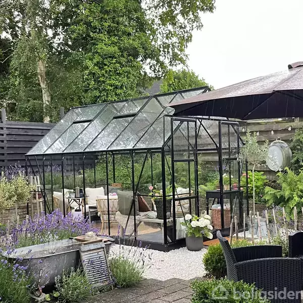 Drivhus med glasvægge står i en frodig have omgivet af grønne planter og lavendel. Indenfor ses stole og bord, skabt til afslapning. Baghaven er indrettet med fliser og træer.