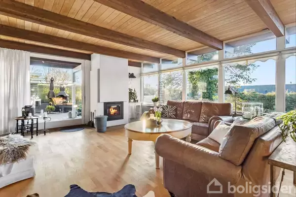 Stue med brændeovn, hvor flammerne brænder, omgivet af store vinduer med udsigt til have. Rummeligt med brune møbler, træloft og lyse trægulve, skaber en lys og hyggelig atmosfære.