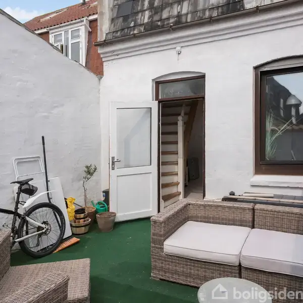 En gårdhave med flettede udendørsmøbler og en cykel står opstillet på en grøn tæppeflade. En åben dør fører ind i huset, omgivet af hvide vægge.