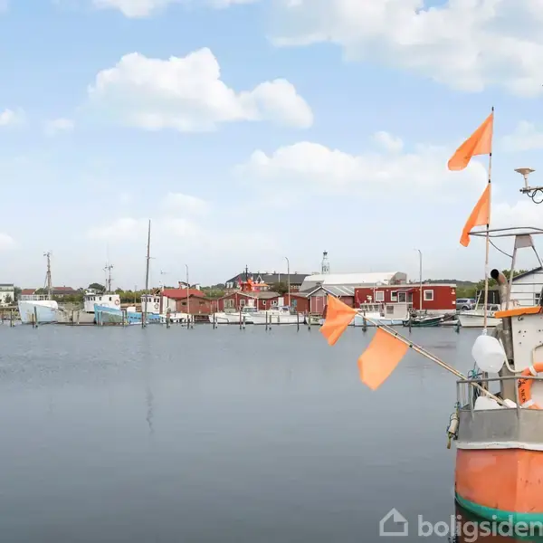 Båd ligger stille i havn med orange flag vejrende. I baggrunden ses flere både, røde bygninger og en rolig vandflade under en blå, lettere skyet himmel.