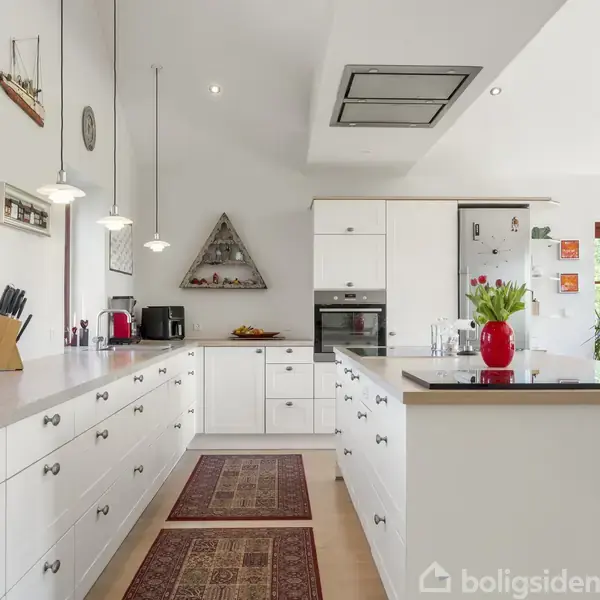 Et lyst, moderne køkken med hvide skabe og sorte greb, har en central ø med en indbygget kogeplade. Dekoreret med planter og røde tulipaner i en vase.