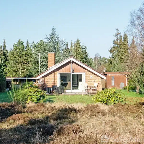Et rødt murstenshus står stille midt i en frodig have omgivet af høje træer. Foran huset findes en terrasse med havemøbler.