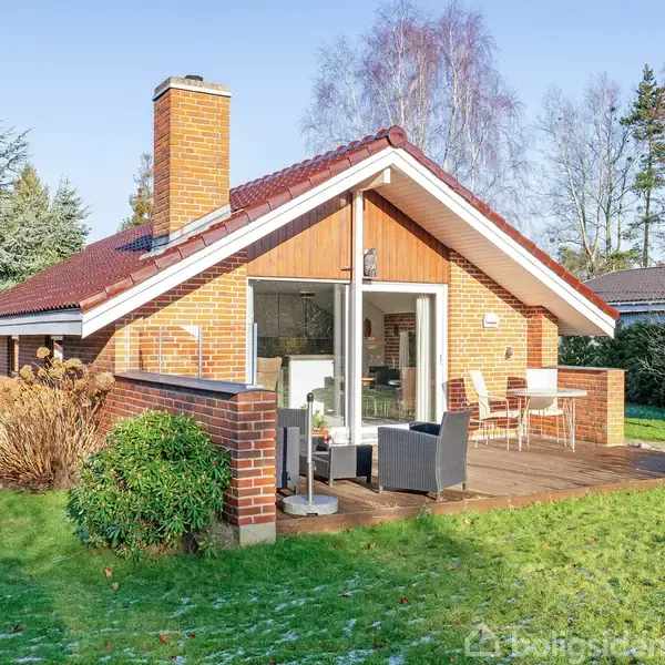 Et rødstenshus med skråt tag står i en grøn have. En terrasse med moderne udendørsmøbler er foran huset. Omkring er der træer og en klar himmel.