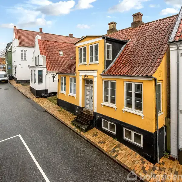 Gult hus står stille langs brostensbelagt gade med røde tage, omkranset af nabohuse og overskyet himmel. En bil er parkeret længere nede ad vejen.