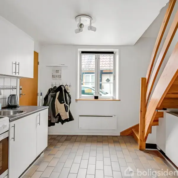 En moderne, funktionel køkken med trætrappe til højre. Står åbne skabe og en sort kogeplade til venstre. Lyse vægge, flisegulv og et stort vindue mod en gårdhave udenfor. Hvidevarer og tøj på knager.