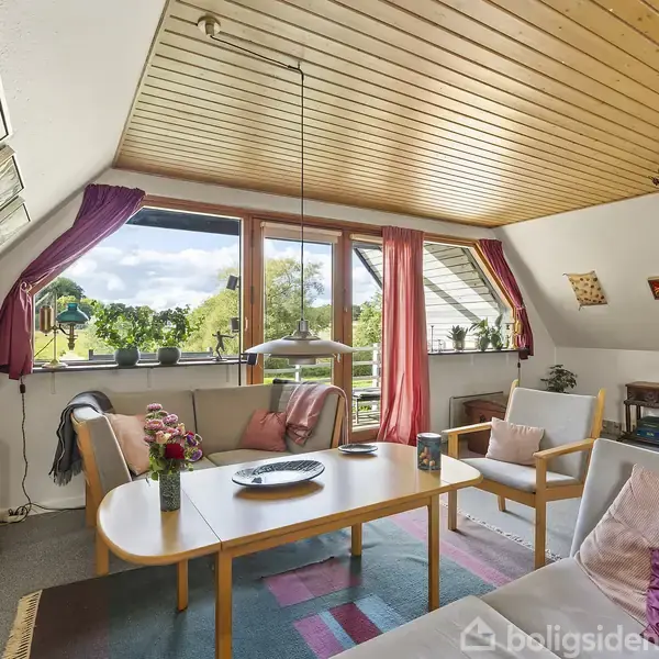 Stue indrettet med sofabord, sofa og lænestole, omgivet af hylder og lamper. Store vinduer med udsigt til træer giver lysindfald. Tagvinduet hælder let, og dekorative puder pynter møblerne.