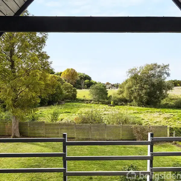 Et terrasselignende område med gelænder indrammer udsigten til en frodig have med træer og marker i baggrunden, omgivet af et hegn.