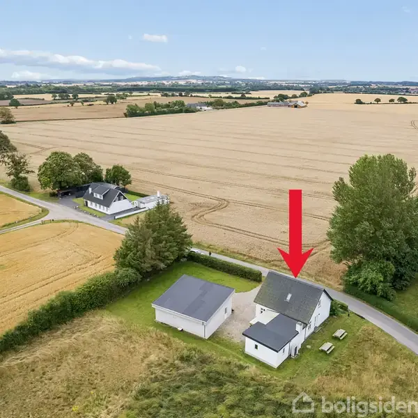 Huset ligger på en stor mark med en rød pil pegende ned på det. Omgivet af grønne træer og marker under en blå himmel med skyer.