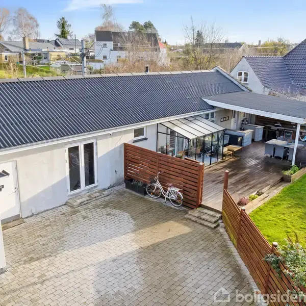 Hus med sort tag ligger ved flisebelagt gårdsplads. To cykler står ved træskur. Terrasse med overdækning ved siden af huset. Grøn græsplæne og flere huse i baggrunden.