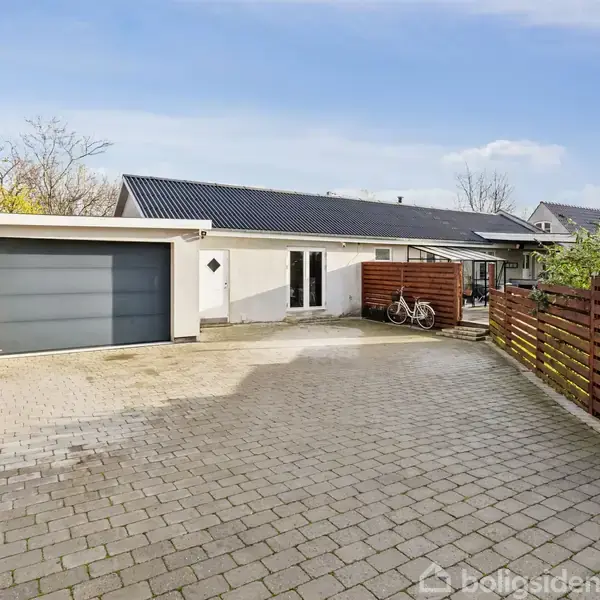 Et moderne hus står stille. Garageporten er lukket, og to cykler er parkeret ved siden af indgangen. Stenbelagt indkørsel og træhegn omkranser huset i et villakvarter.