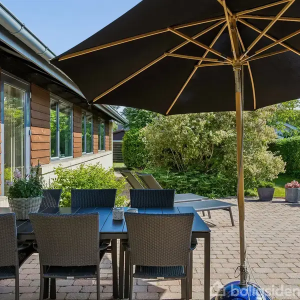 Terrassebord med seks stole står under en stor parasol. Det hele er placeret på en flisebelagt terrasse foran et hus. Baggrunden er grøn med buske og blomsterkrukker.
