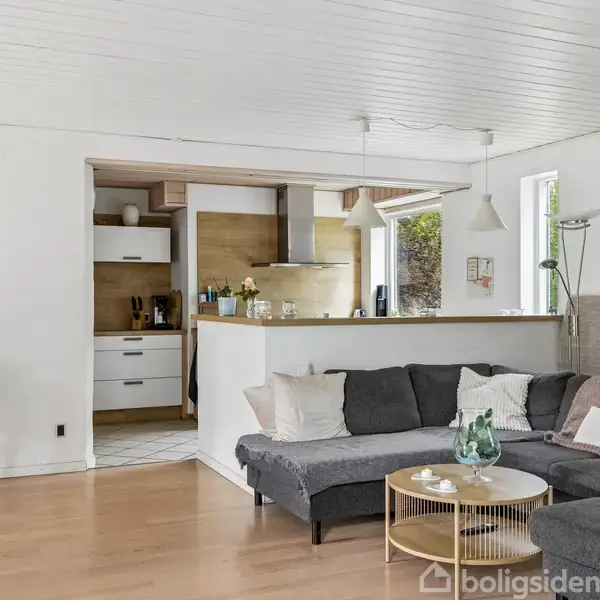 Grå sofa står i lyse stue med træbord. I baggrunden er et åbent køkken med hvidt skab, emhætte og planter. Store vinduer giver naturligt lys.