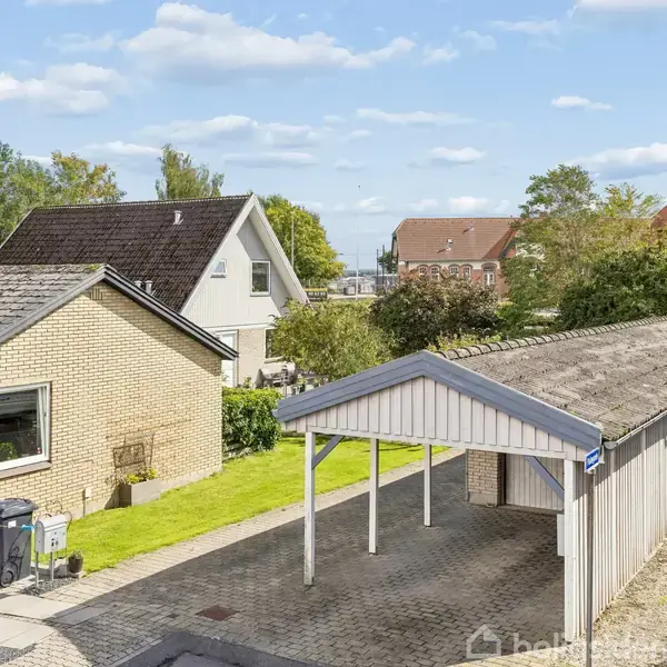 Carport står tom ved siden af et gult murstenshus med græsplæne. Flere huse omgiver området, og en blå skraldespand ses nær husets væg. Himlen er klar med skyer.