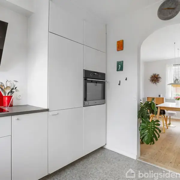 Køkken med hvide skabe og indbygget ovn, rød backsplash ved siden af sort emhætte. I baggrunden ses et spisebord med stole og en grøn plante.