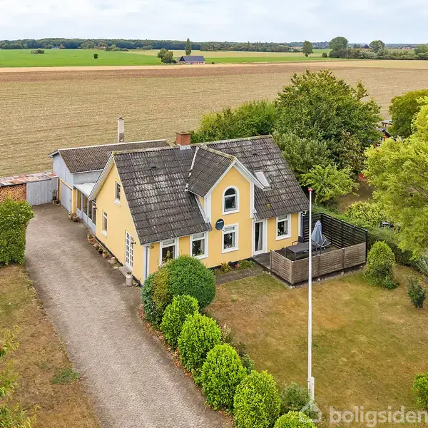Et gult hus med gråt tag står omgivet af en have med grønne buske og træer. En grusvej fører til huset, og det ligger i et åbent landskabsområde med marker.