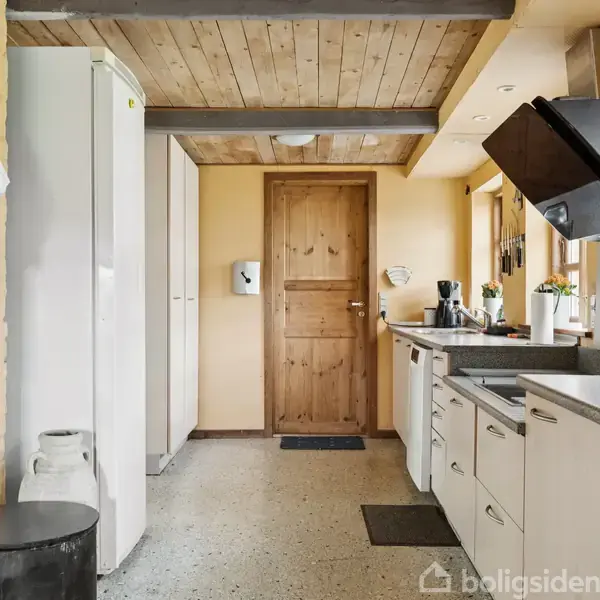 Køkken med hvidt inventar og gråt bordbord, hvor køleskab og sorte apparater er synlige. Trædør i baggrunden, lyse vægge og loft med træbjælker.