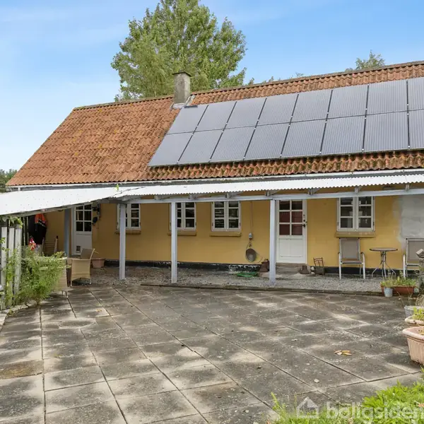 Et gulmalet hus med rødt tegltag har solpaneler monteret. Handlingen indbefatter en overdækket terrasse med stole og borde. I baggrunden ses grønne træer og buske.