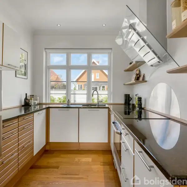 Køkken med moderne apparater og trægulv. Skabe og hylder er fyldt med diverse køkkengenstande. Et vindue giver udsigt til et solrigt udendørsområde med huse i baggrunden.