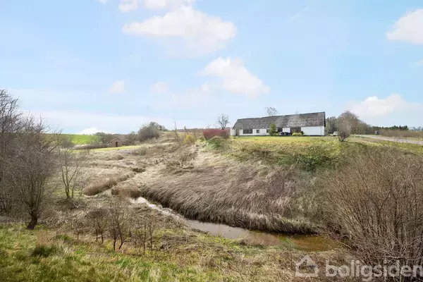 Et hvidt hus med stråtag står på en bakke. Omkring huset ses åbne marker og en lille bæk snor sig gennem landskabet under en blå himmel med skyer.