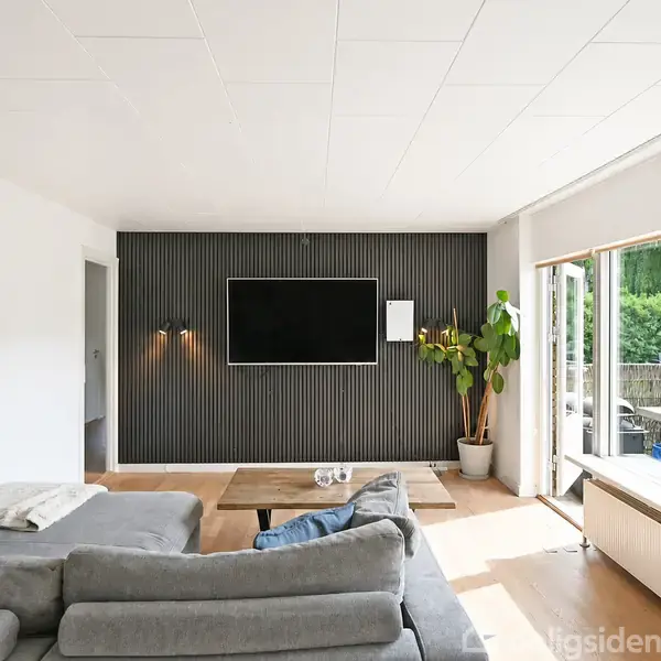 En grå sofa står i midten af en stue med en fladskærm på en sort væg. Store vinduer åbner mod en terrasse med grill og planter udenfor.