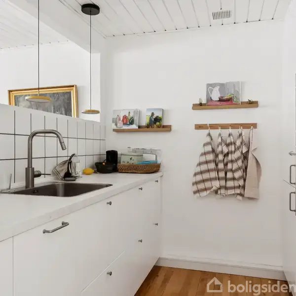 Køkken med hvide skabe og mørk bordplade. En vask med høj hane, flisevæg, og ovne indbygget i højre væg. Trægulv og hylder med dekorative genstande.