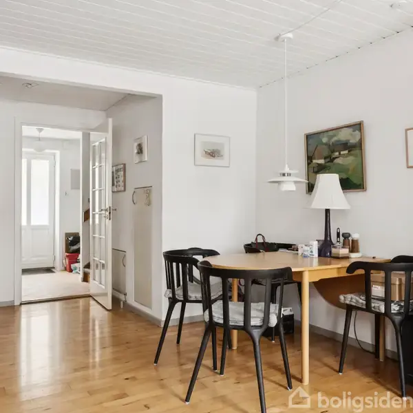 Et spisebord med fire sorte stole står i et lyst rum med parketgulv. To lamper hænger over bordet, og væggene er dekoreret med billeder. En døråbning fører til en gang.