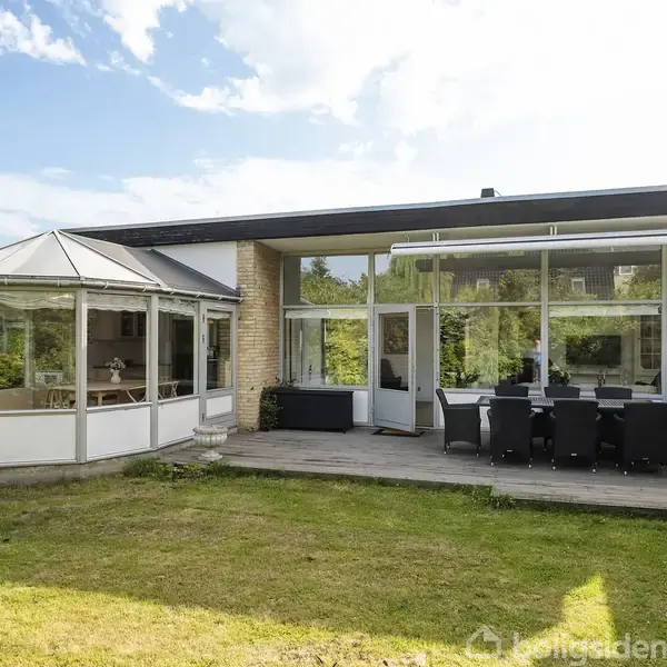 Et moderne hus med store vinduer og en udestue ligger i en have med græsplæne. En udendørs terrasse har et bord med otte stole.