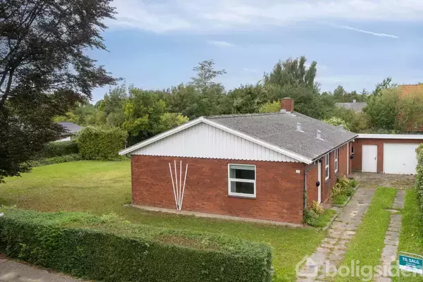 Et rødt murstenshus med hvide detaljer står i en have med en plænesti. En garage ses i baggrunden. Et skilt foran huset viser teksten "TIL SALG".