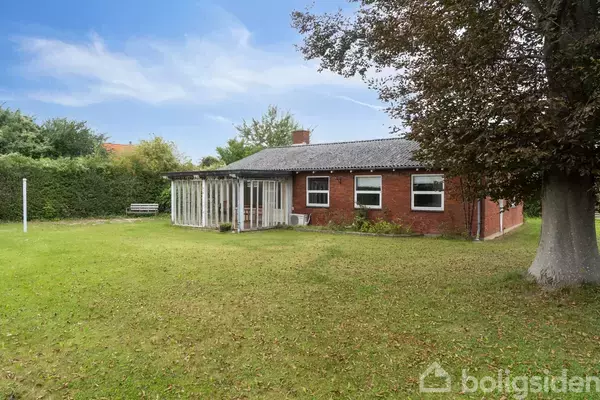 En rød murstensvilla med udestue står stille i en frodig have med græsplæne og træer, omgivet af tæt vegetation og en blå himmel i baggrunden.