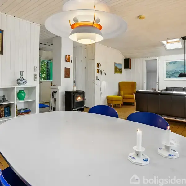 Et lyst spiseområde med et ovalt bord omgivet af blå stole. To tændte stearinlys står på bordet. I baggrunden er en stue med sofasæt og en pejs.