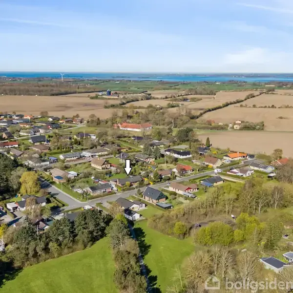 Et fugleperspektiv af en landsby med huse og veje omgivet af marker og skove. I det fjerne ses en kystlinje med blå himmel og vand. En pil markerer et bestemt hus.
