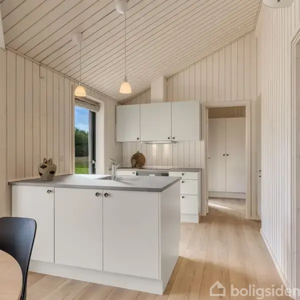 Et lyst køkken med hvide skabe og en central ø, oplyst af pendellamper. En dør fører til et rum med sengebelysning. Trævægge og -loft skaber en hyggelig atmosfære.