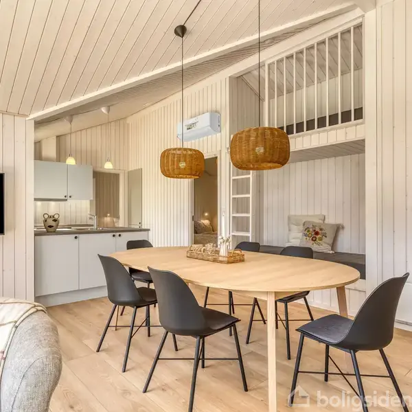 Rundt spisebord med seks stole står i et lyst, moderne træbeklædt rum. To flettede pendellamper hænger over bordet. Bagved ses et køkkenområde og en hems med stige.