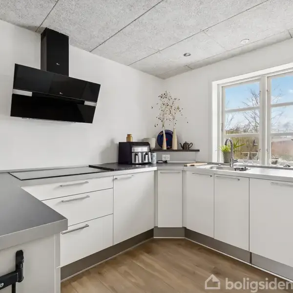 Et moderne køkken med hvide skabe og sort emhætte. Bøtter og planter står i vindueskarmen. Trægulve dækker gulvet under blødt naturligt lys fra et stort vindue.