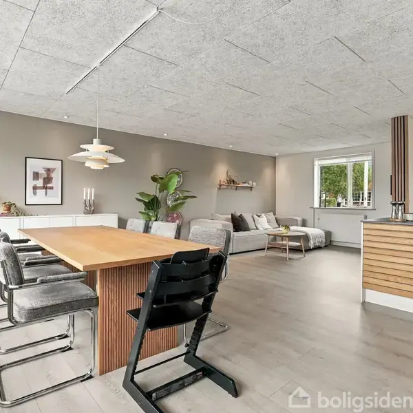 Spisebord med stole står i et moderne, åbent køkken-alrum. Stuen i baggrunden har en sofa, planter og vægdekorationer. Store vinduer giver naturligt lys og udsigt til træer.