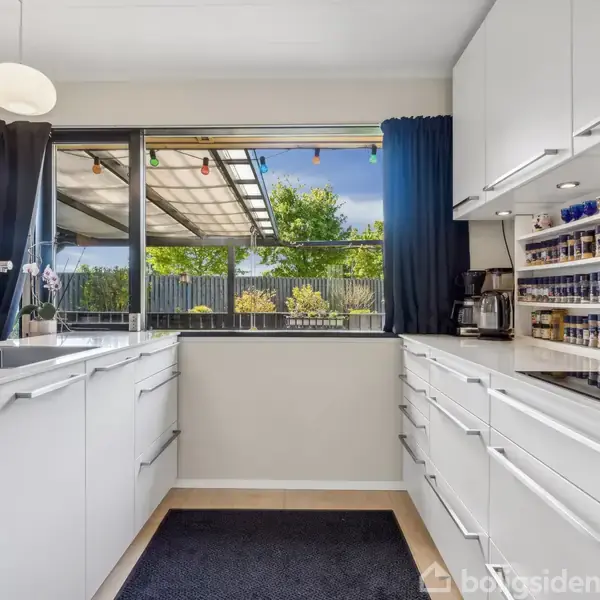 Et moderne køkken med hvide skabe og skuffer omkring en sort bordplade. Køkkenet har en vask, kaffemaskine og krydderihylde. Store vinduer med udsigt til en have fungerer som baggrund.