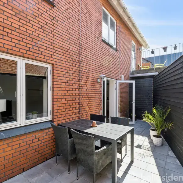 En gårdhave med et sort bord og fire stole står på en flisebelagt terrasse. Det er omgivet af røde murstensvægge og sorte træhegn, med en plante i en hvid potte.