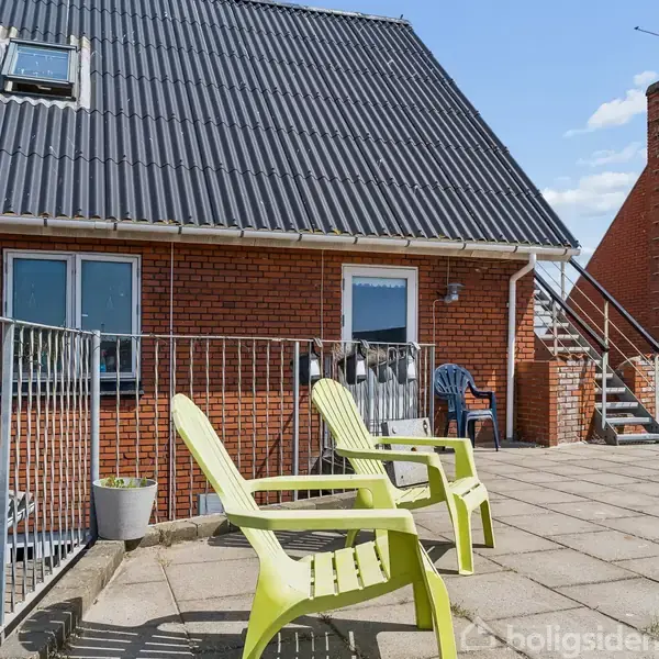 To grønne plastikstole står på en flisebelagt terrasse ved et rødt murstenshus med sort tag. Området er indhegnet med metalgelænder, og der er en udendørs trappe.