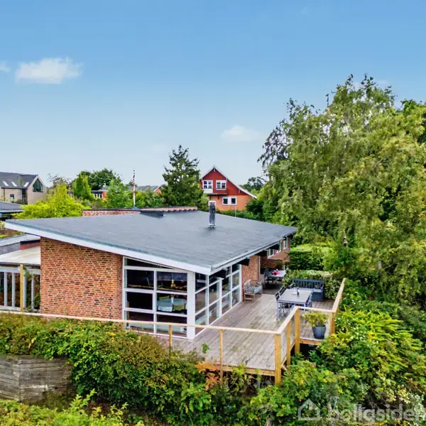 Huset, en moderne rød murstensbygning med fladt tag, står på en forhøjning omgivet af frodig grønt. I baggrunden ses flere villaer og en blå himmel.